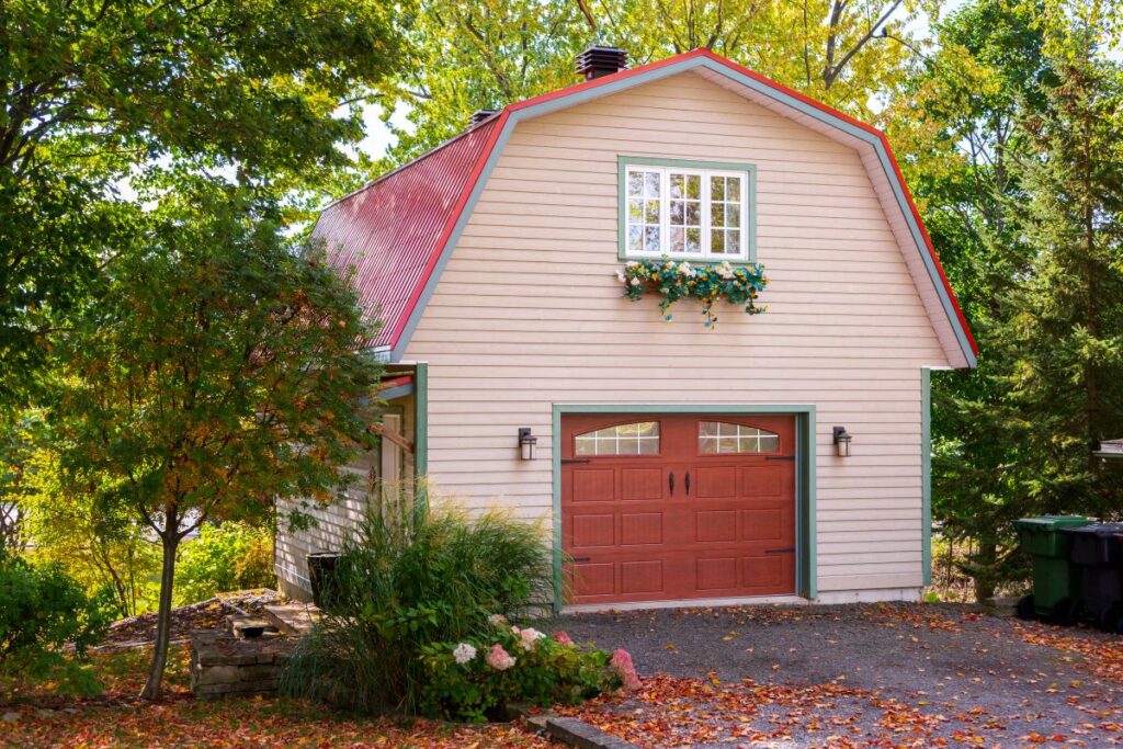 A cream-colored barn-style garage with a gambrel roof, red door, green trim, and a window box of flowers, surrounded by trees and autumn leaves on the ground.
