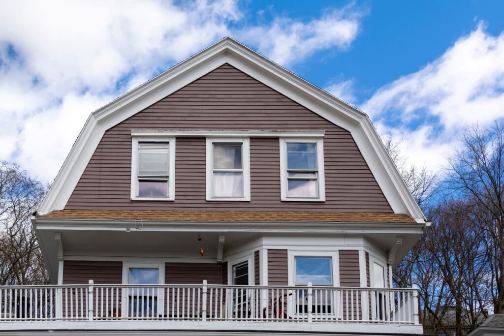 A two-story house with brown siding, white trim, and a gambrel roof features a front porch with a white railing, set against a blue sky with scattered clouds and leafless trees.