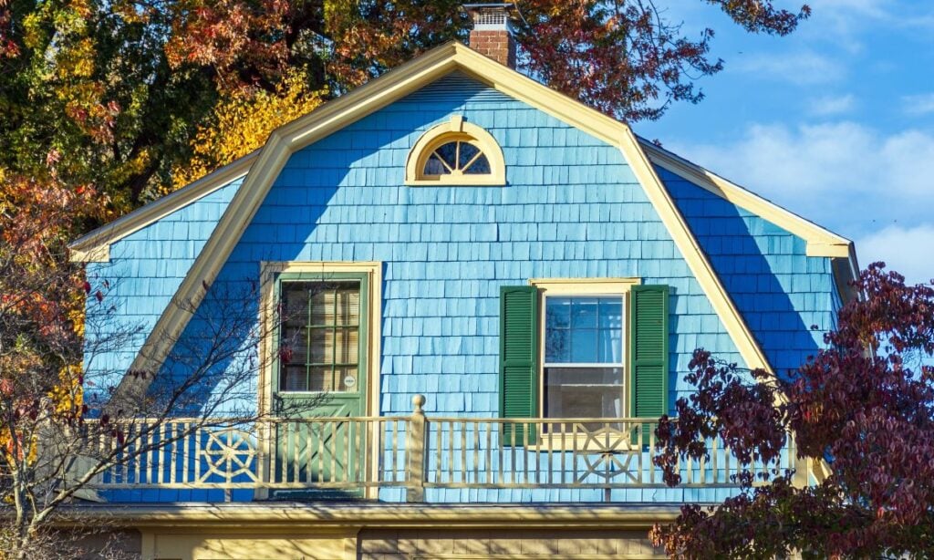 A blue house with cream trim, green shutters, and a Dutch-style gambrel roof is nestled among autumn trees. A small balcony with a decorative railing adds charm to this inviting home.