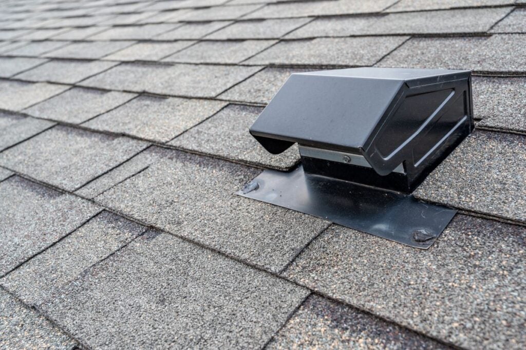 A close-up view of a black roof vent cap installed on gray asphalt shingles, highlighting the textured roof surface and the sleek design of the vent.