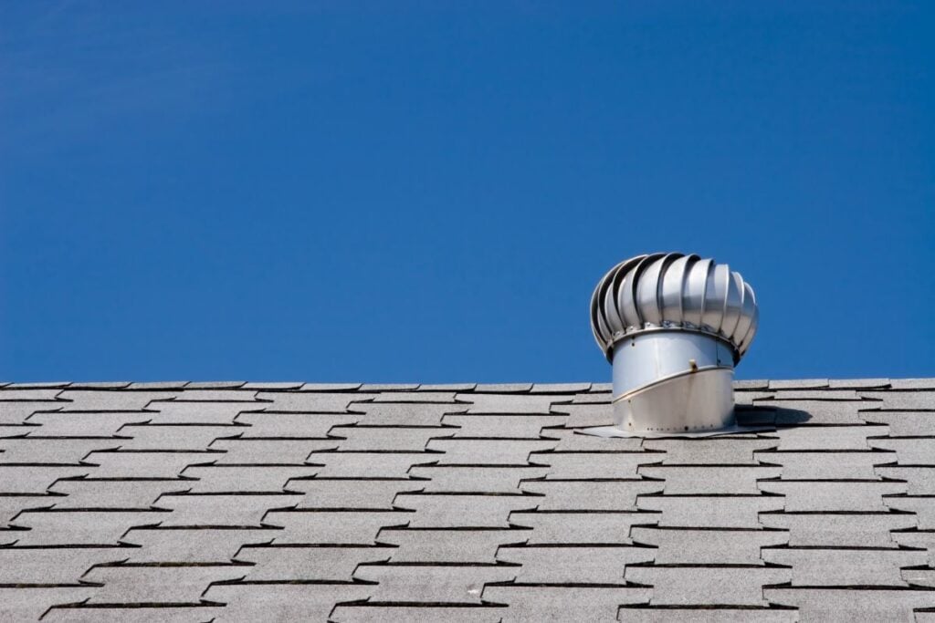 A metal turbine roof vent cap is installed on a gray shingled roof under a clear blue sky.
