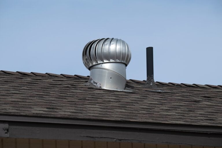 A silver turbine roof vent and a black metal pipe protrude from the brown shingle roof of a building under a clear blue sky.