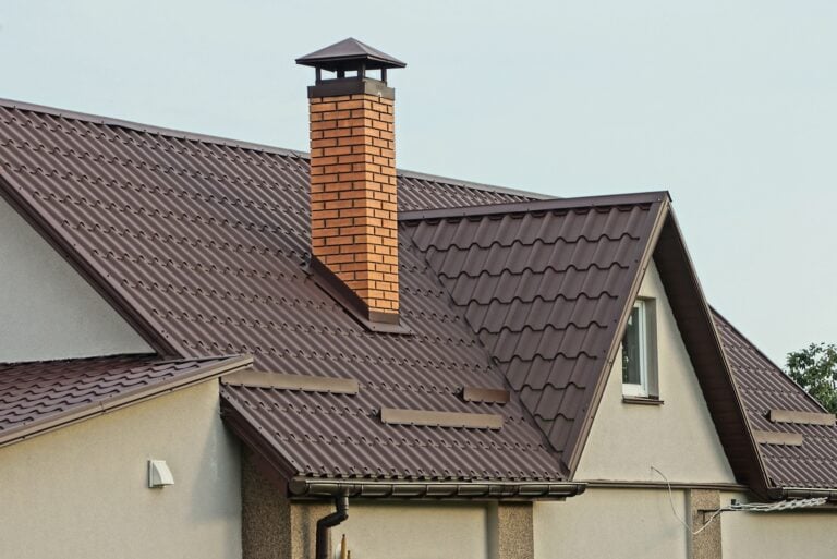 A house with light-colored walls and a brown metal tiled roof, featuring a prominent red brick chimney and several roof angles, with a small window visible under the gable.