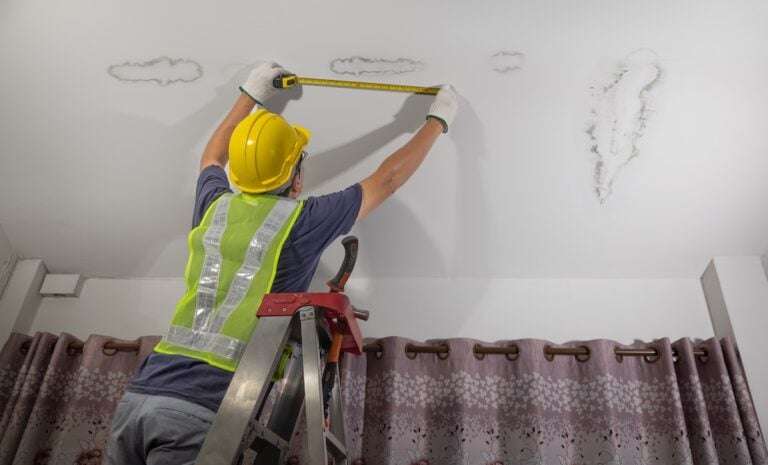 A worker in a yellow hard hat and reflective vest stands on a ladder, carefully measuring water spots on the ceiling but no leak can be seen above a set of purple curtains.