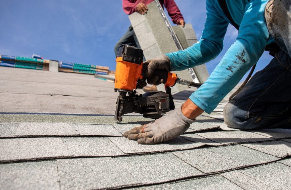 A worker wearing gloves and a blue shirt uses a nail gun to secure asphalt shingles to a roof, helping prevent nail pops on roof surfaces, while another person in the background carries more shingles under a clear blue sky. nail pops on roof