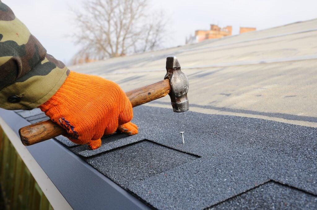 A person wearing an orange glove uses a hammer to drive a nail into black asphalt roof shingles on a sloped roof, helping prevent nail pops on the roof.