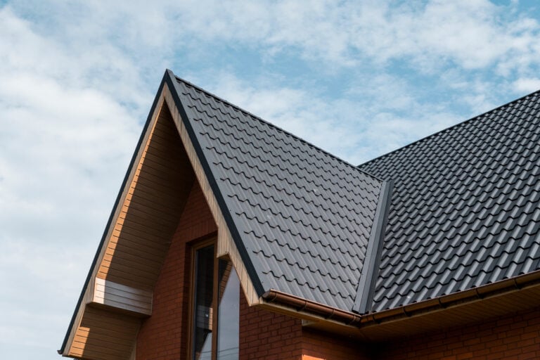 A close-up view of a modern house roof featuring a dark gray metal roof coating, brick walls, and wooden accents, set against a blue sky with scattered clouds.