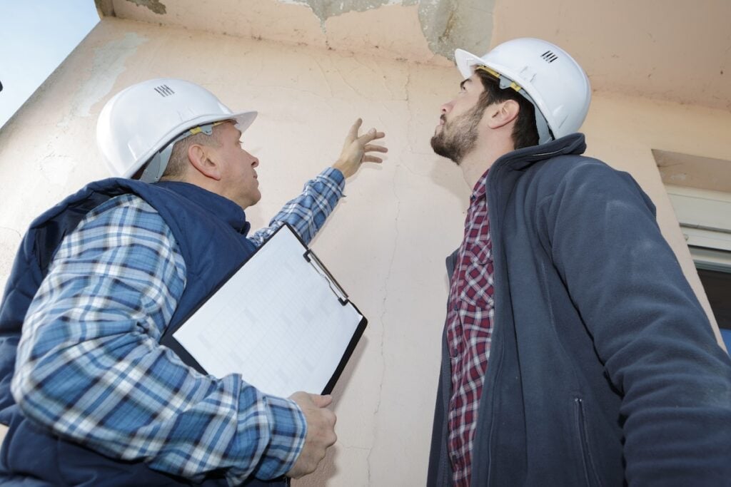 Two construction workers wearing hard hats examine cracks and damage on a building wall. One worker points at the damage, discussing possible causes like water spots on ceiling but no leak, while the other looks up, holding a clipboard.