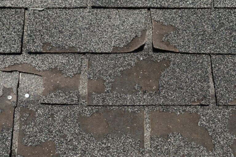 Close-up of weathered, damaged roof shingles with missing roofing granules, curled edges, and exposed nails, indicating significant wear and potential need for repair or replacement.