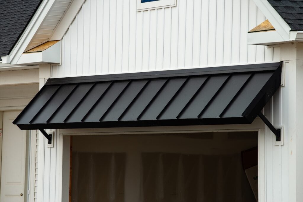 A black metal awning is installed above the entrance of a white building with vertical siding. The area beneath the awning appears to be an unfinished garage with exposed drywall.