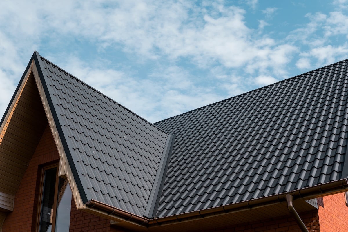 A close-up view of a modern house with a metal roof that looks like shingles, dark gray tiles, brown brick walls, and a clear blue sky with scattered clouds in the background.