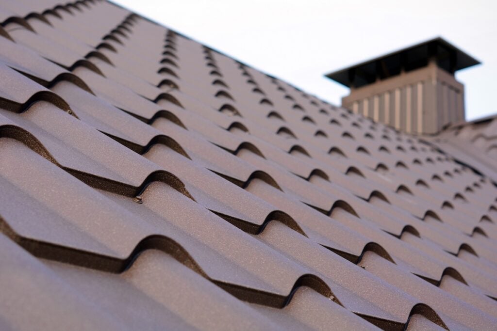 Close-up view of a metal roof that looks like shingles, with a chimney vent in the background, highlighting the wavy pattern and texture of the brown roofing material.