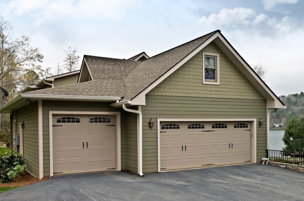 Light green house with beige trim showcases attractive siding colors, featuring two garage doors—one single and one double—on a paved driveway. A window sits above the double garage, with trees and a lake visible under a partly cloudy sky.