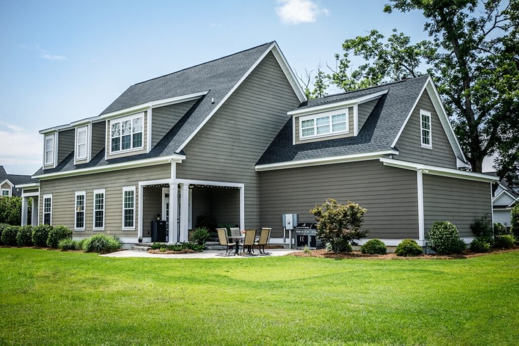 A modern two-story gray house with white trim, large windows, and a covered patio area. The backyard features a neatly mowed lawn, shrubs, and an outdoor dining set—showcasing one of today’s most popular siding colors.