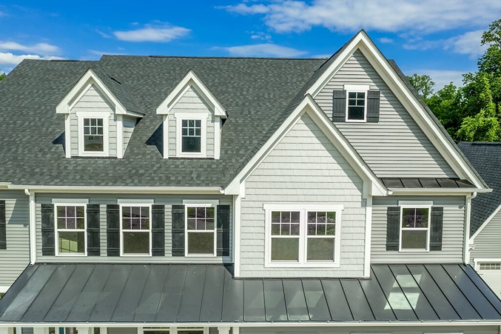 Front view of a modern gray house with white trim, multiple windows, black shutters, and a metal roof over the porch highlights stylish siding colors against clear skies in the background.