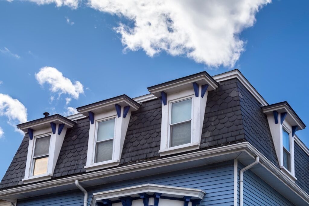 The image shows the upper part of a blue house with a mansard roof featuring three dormer windows, white trim, and decorative blue accents, set against a partly cloudy sky.