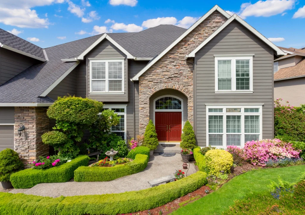 Two-story suburban house with stylish siding colors, gray siding, stone accents, and a red double front door. The yard features manicured shrubs, colorful flowers, and a curved walkway leading to the entrance under a blue sky with clouds.