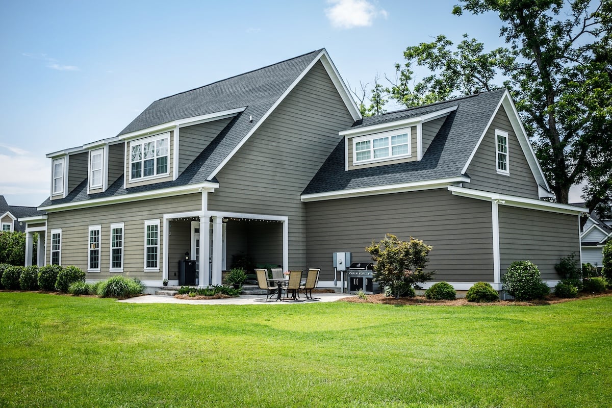 A modern gray two-story house with white trim, large windows, and a covered patio. Durable roofing granules enhance the roof’s longevity. The patio set, grill, and lush green lawn with shrubs and small trees create a welcoming outdoor space.