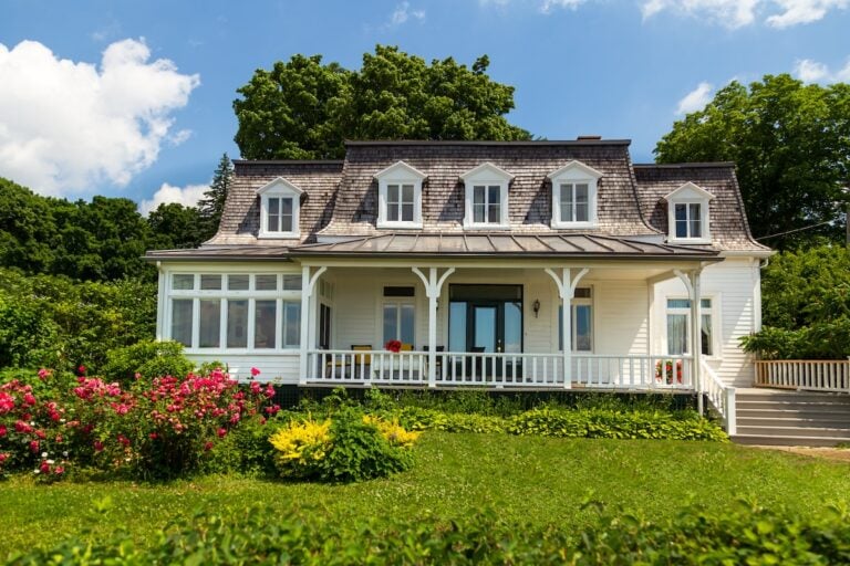 A charming white house with a large porch, dormer windows, and a manicured lawn surrounded by green trees and colorful flowering bushes under a blue sky with scattered clouds.