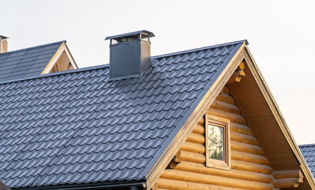 A close-up of a log cabin roof with a metal roof that looks like shingles, featuring a chimney and a window reflecting trees, all set against a light sky.
