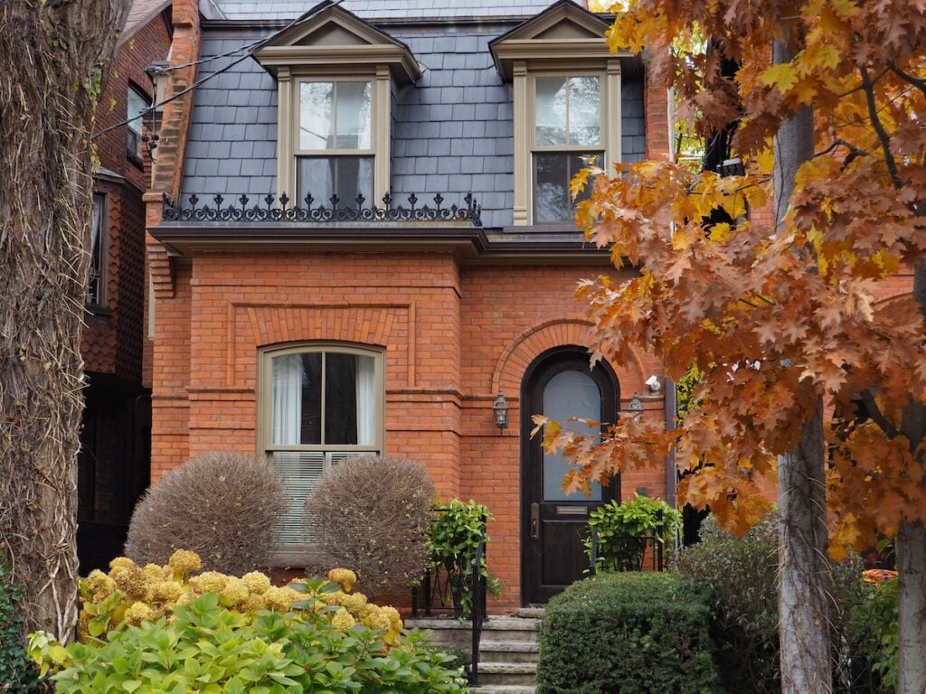 A two-story red brick house with two dormer windows, arched front door, manicured shrubs, and autumn trees with orange leaves in the foreground.