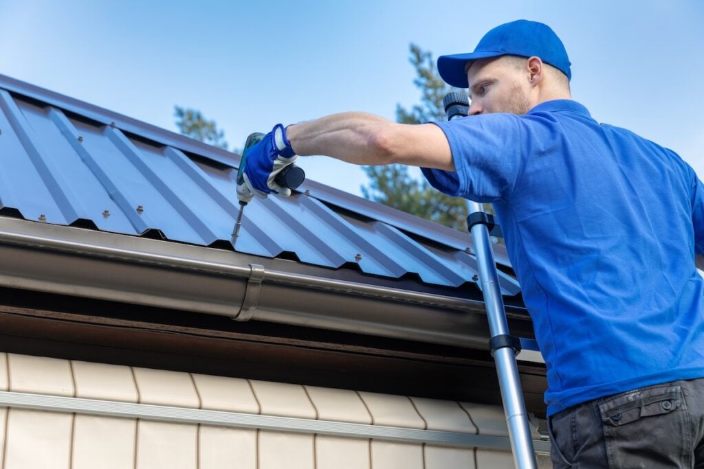 A worker in a blue uniform and cap stands on a ladder, using a power drill to secure a metal roof panel on a building. Trees and blue sky are visible in the background.