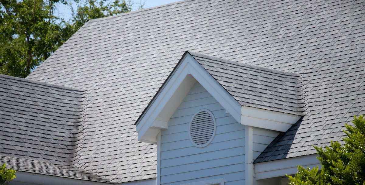 Close-up of a house roof with gray asphalt shingles showing roofing granules, light blue siding, and white trim. A small gable with a circular vent is featured in the center, framed by green trees in the background.