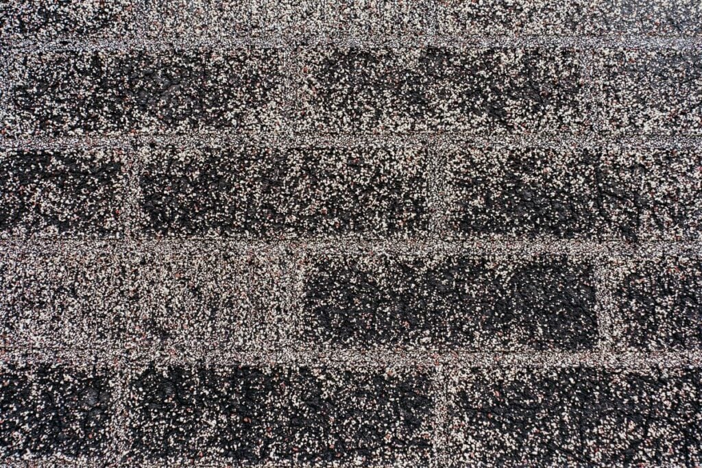 Close-up of dark asphalt roof shingles with a rough, textured surface and visible roofing granules, arranged in a staggered brick-like pattern. The shingles display a speckled look from the mix of light and dark granules.