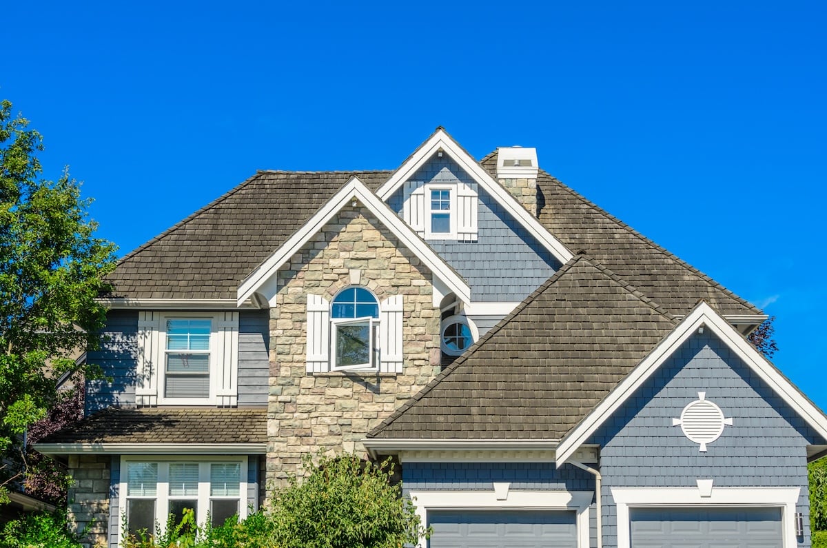 Two-story suburban house with gray-blue siding, showcasing popular types of siding, complemented by stone accents, white shutters, and a gabled roof under a clear blue sky, partially surrounded by green shrubs and trees.