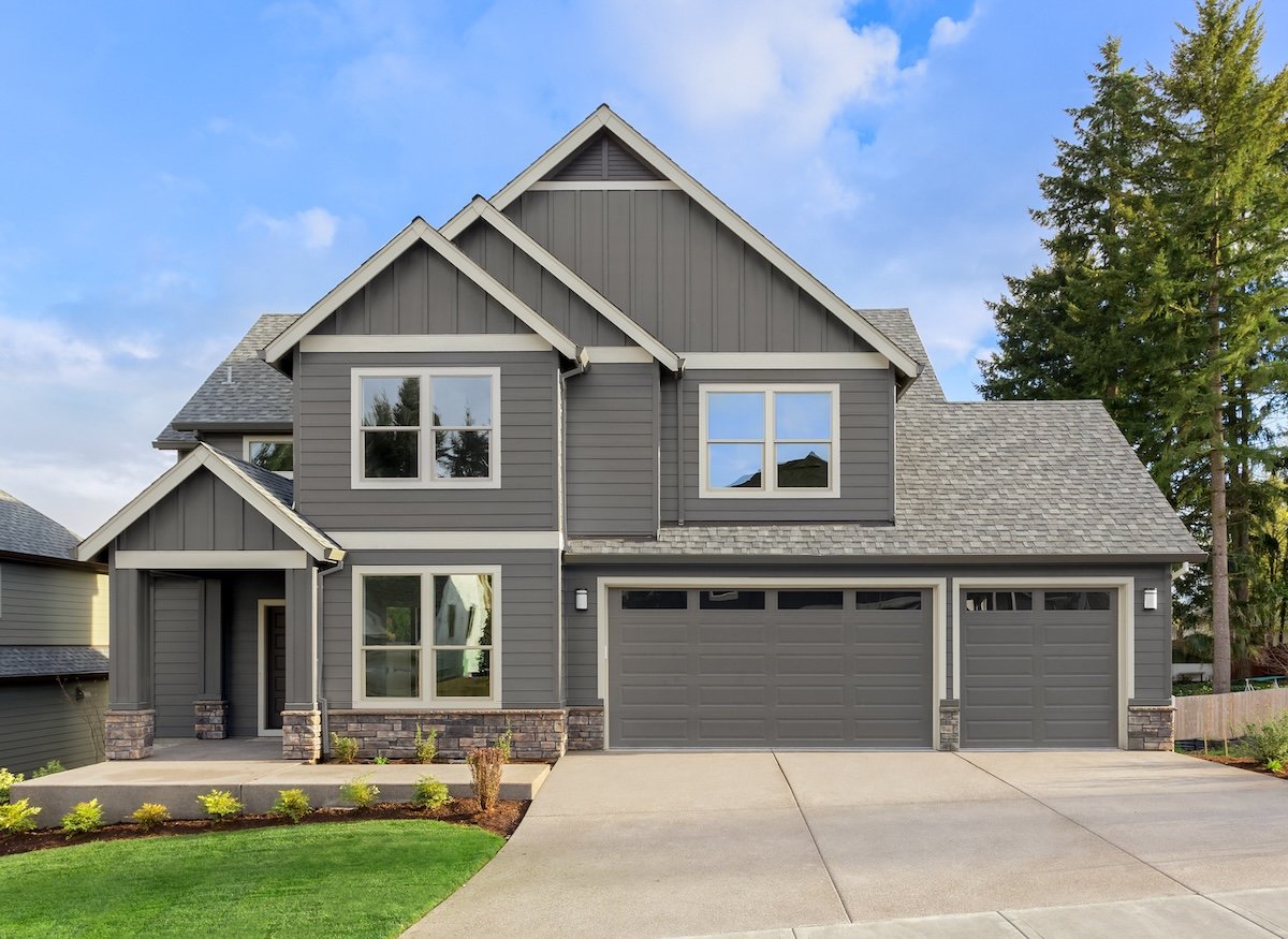 A modern two-story gray house with white trim, large windows, and a three-car garage features expert craftsmanship by a Virginia roofing company, surrounded by manicured shrubs and tall trees under a partly cloudy sky.