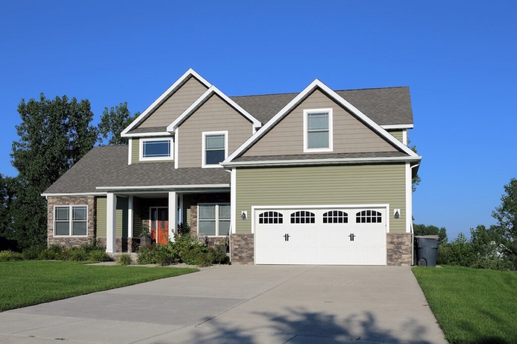 This two-story suburban house features a green and beige exterior with white trim, a double garage door, covered front porch, and large driveway, all beautifully maintained by a trusted Virginia roofing company.
