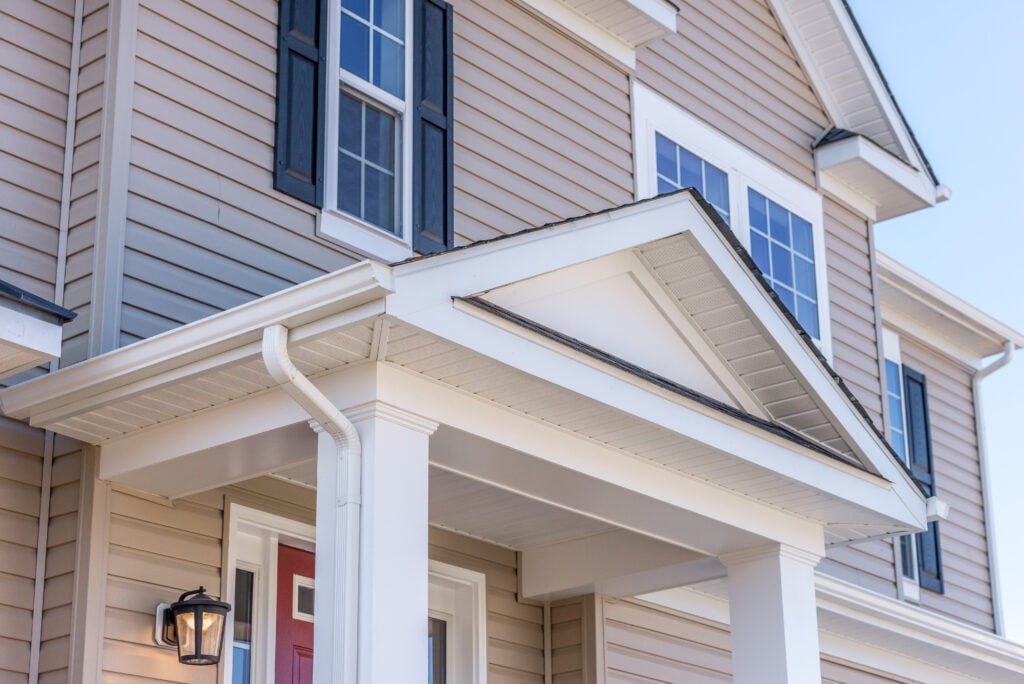Close-up of the front exterior of a modern house featuring the best siding for house durability—tan siding, white trim, columns, a red door, black shutters, and a porch light beside the entrance.