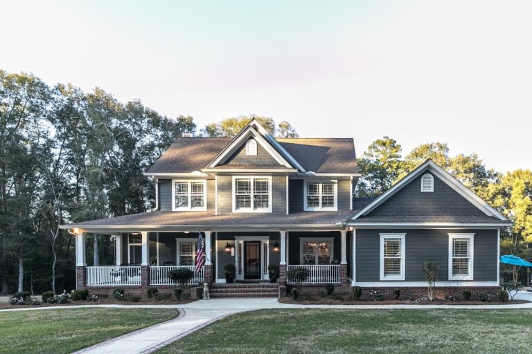 Two-story gray house with white trim, featuring the best siding for house durability, a wraparound porch, and an American flag by the entrance, surrounded by trees and a lawn with a curved walkway leading to the front steps.