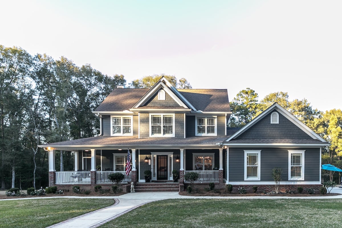 Two-story gray house with white trim, featuring the best siding for house durability, a wraparound porch, and an American flag by the entrance, surrounded by trees and a lawn with a curved walkway leading to the front steps.