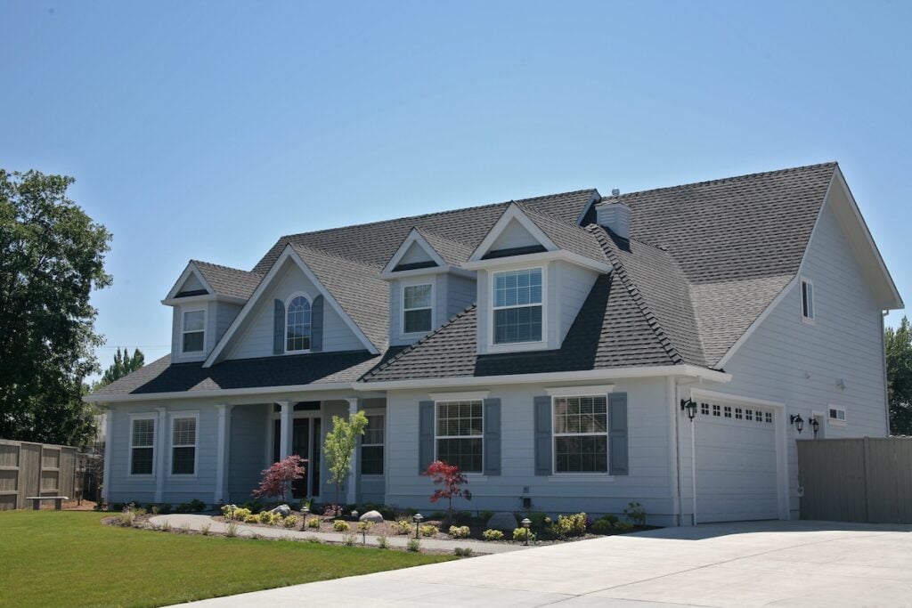 A large two-story suburban house with the best siding for house in gray, white trim, multiple gables, and a two-car garage. The front yard features small shrubs and trees, with a wide driveway leading to the garage.