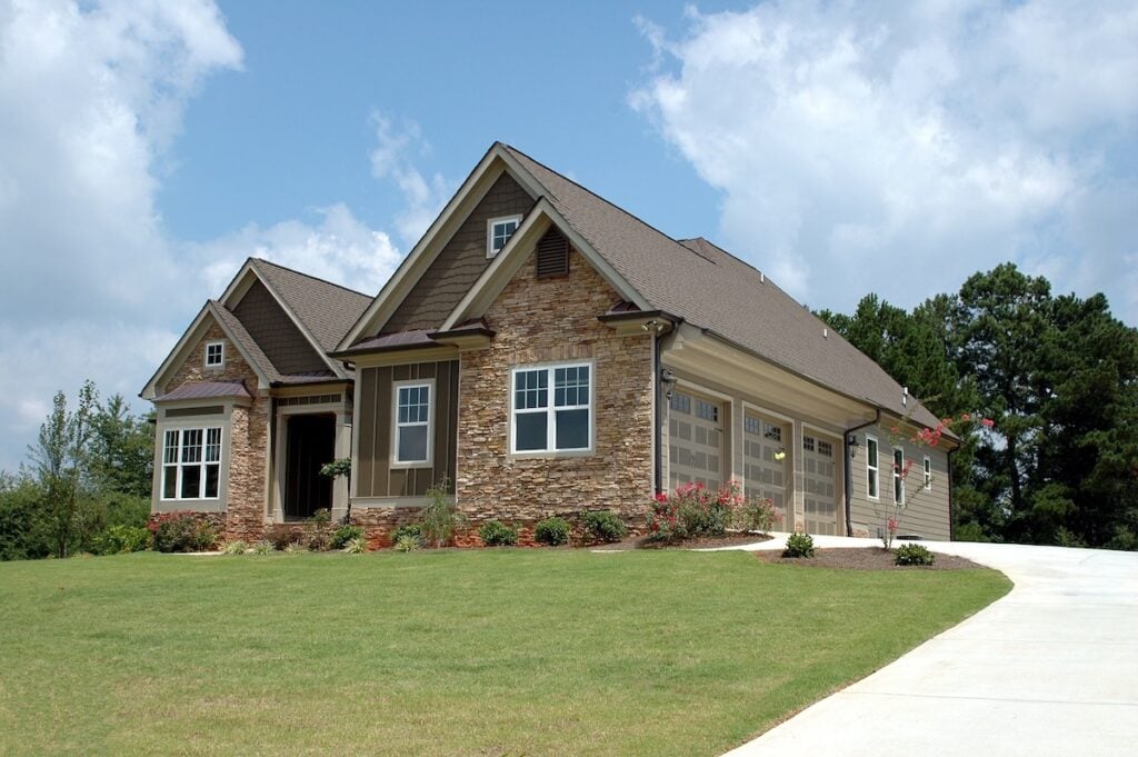 A single-story suburban house with stone and best siding for house exterior, large windows, a three-car garage, and a well-maintained front yard with green grass and flower beds, under a partly cloudy sky.
