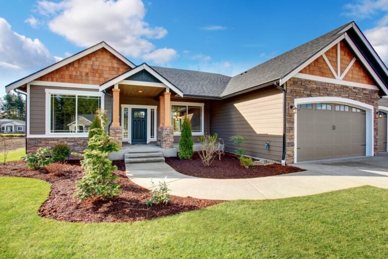Single-story suburban house featuring brown siding materials and stone accents, a covered front porch, attached two-car garage, landscaped front yard with shrubs, and a concrete driveway on a sunny day.