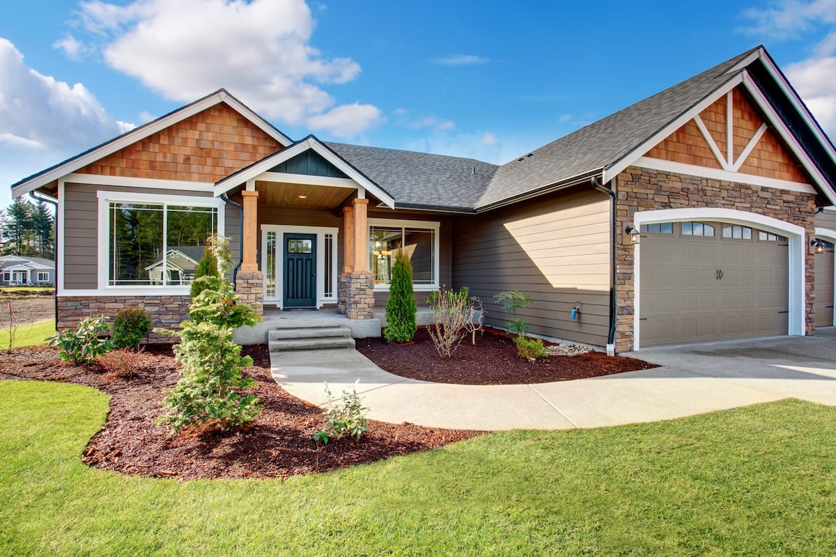 Single-story suburban house featuring brown siding materials and stone accents, a covered front porch, attached two-car garage, landscaped front yard with shrubs, and a concrete driveway on a sunny day.