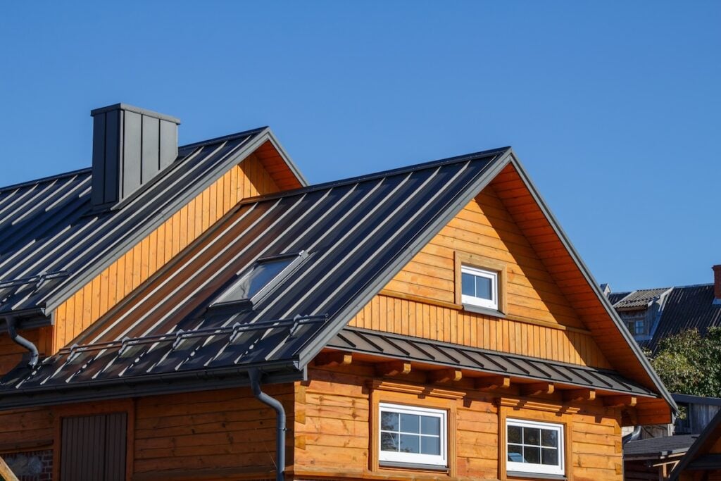 A wooden house with a steep metal roof and small windows showcases classic siding materials, set against a clear blue sky on a sunny day.