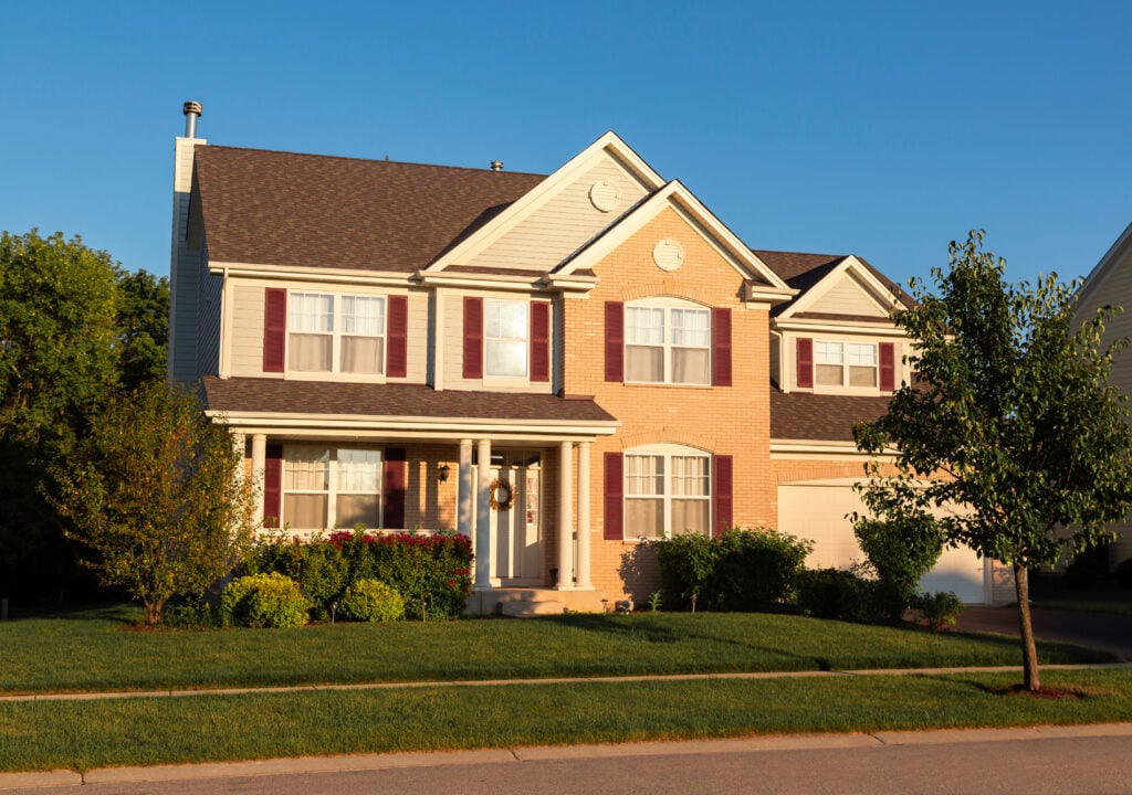 Two-story suburban house featuring quality siding materials with beige siding and brick exterior, white trim, and dark shutters. Includes a covered front porch, two-car garage, and is set amid trees and a well-kept lawn under a clear blue sky.