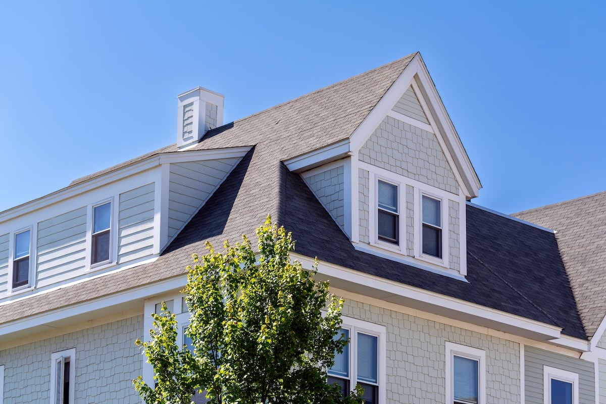 A light gray house with white trim, featuring multiple gables, dormer windows, and a steep shingled roof by a leading Virginia roofing company, is partially obscured by a leafy green tree under a clear blue sky.