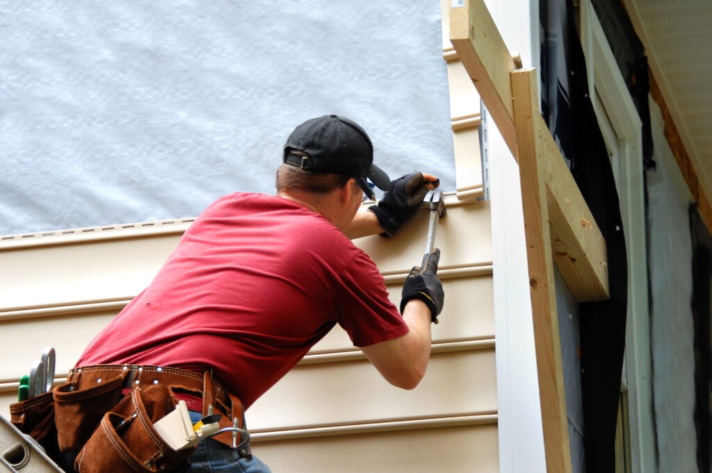 A construction worker in a red shirt and black cap installs beige siding on a house, using a hammer and nail. He wears a tool belt with various tools and stands close to an unfinished section of the building.