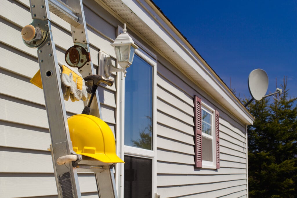 A ladder leaning against a beige house with a yellow hard hat, gloves, and a hammer hanging on it. The house has white trim, a window with shutters, a wall lantern, and a satellite dish. Trees and blue sky are in the background.