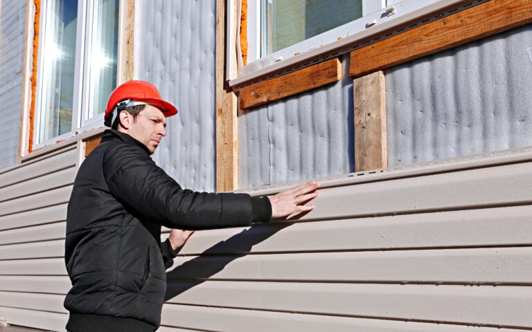 A man wearing a red hard hat and black jacket installs beige vinyl siding on the exterior wall of a building under construction.