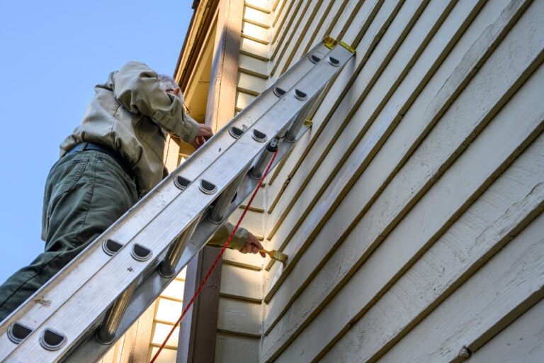 A person standing on a ladder paints the exterior wall of a house with a paintbrush, reaching up towards the trim near the roof on a clear, sunny day.