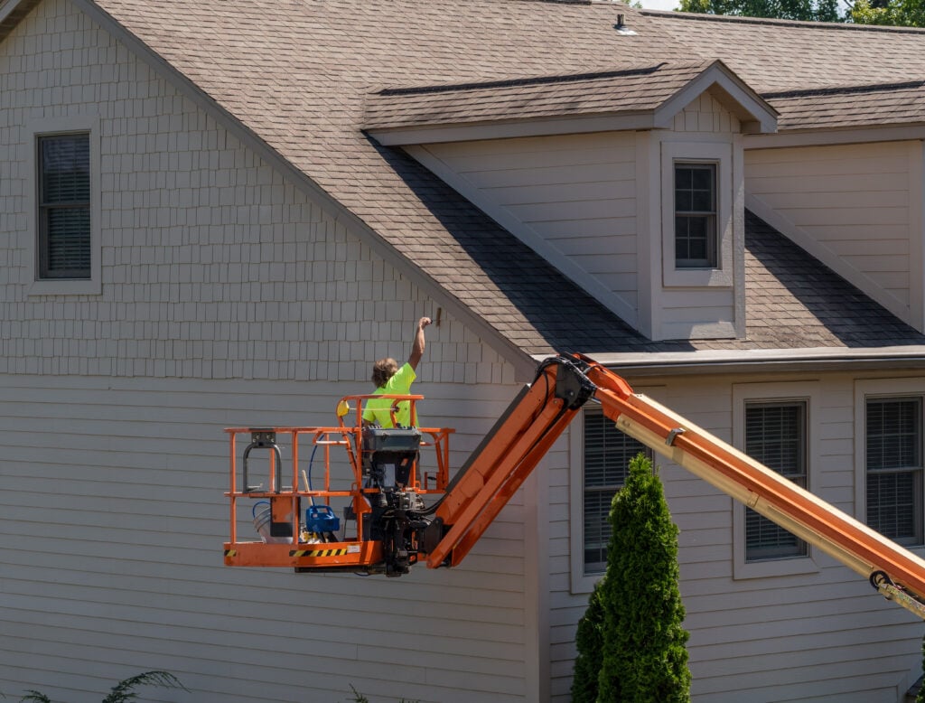 A worker in a neon shirt stands on an orange boom lift, painting the upper exterior wall of a beige house with gray trim on a sunny day. can you paint vinyl siding