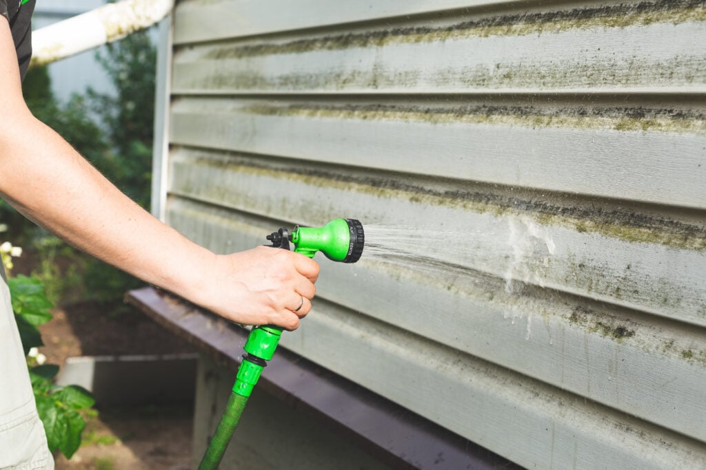A person demonstrates how to clean vinyl siding by using a green garden hose with a spray nozzle to wash dirt and mold from the exterior of a house.