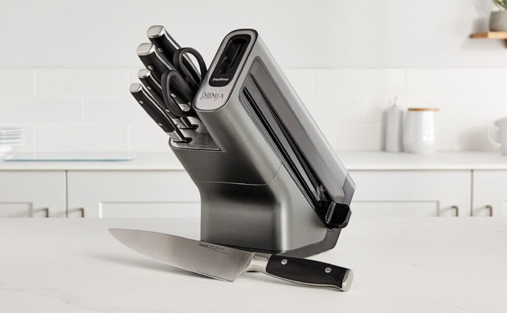 A modern knife block set with several knives and kitchen shears stands on a white kitchen counter, with one large chef’s knife lying in front of it. The kitchen background is bright and minimal.