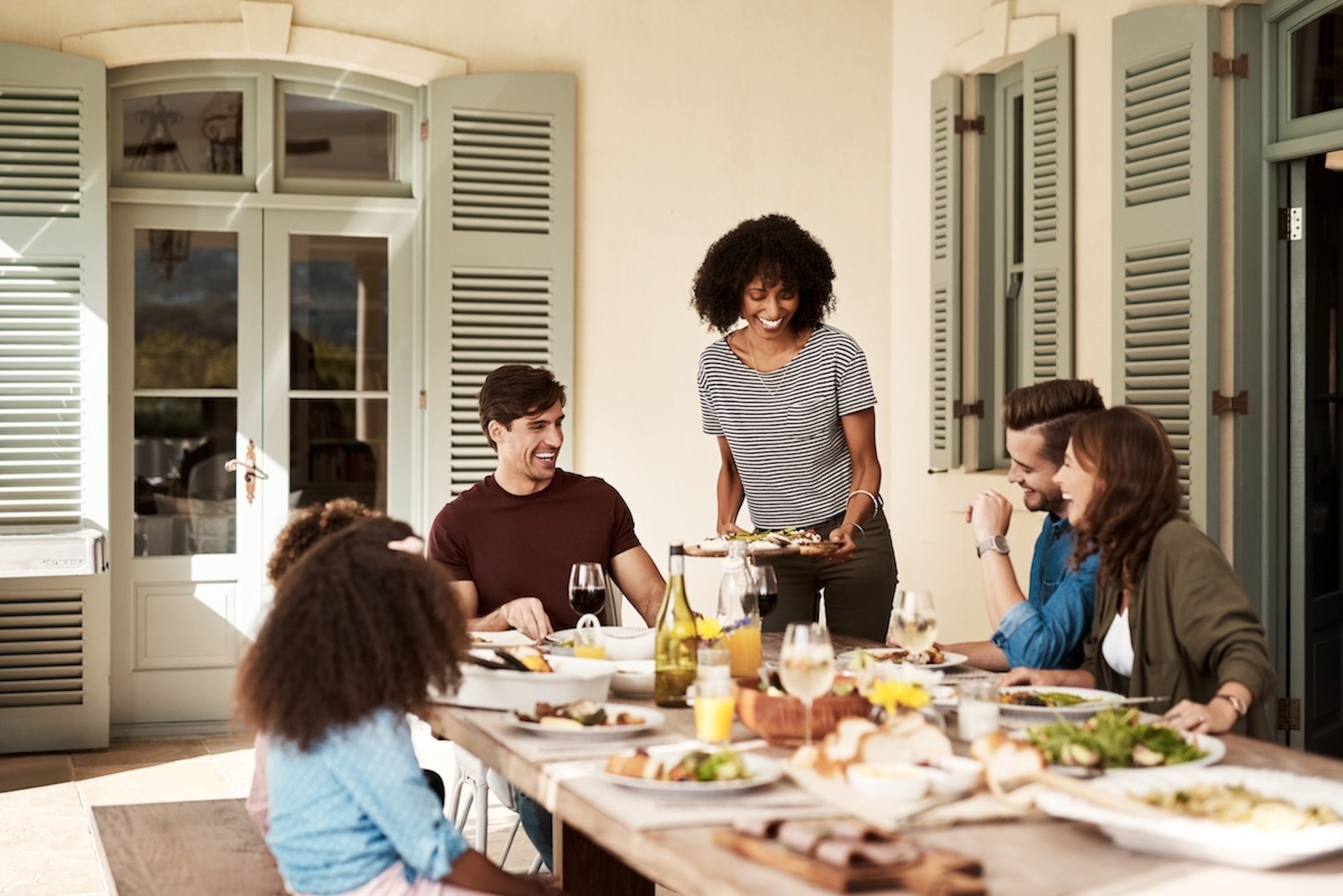 A group of people sitting around an outdoor table, enjoying a meal together. One person is standing, serving food, while others smile and talk. The setting is bright with light green shutters and large windows in the background.