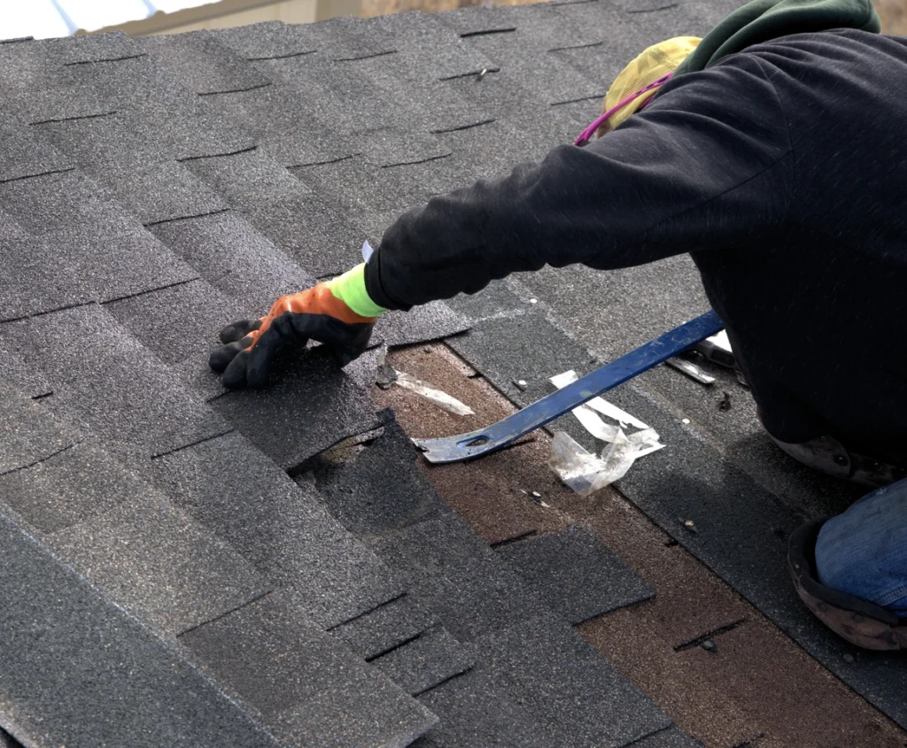A person wearing gloves and a hat uses a crowbar to repair or replace shingles on a sloped roof, showcasing expert roofing Mount Vernon services.